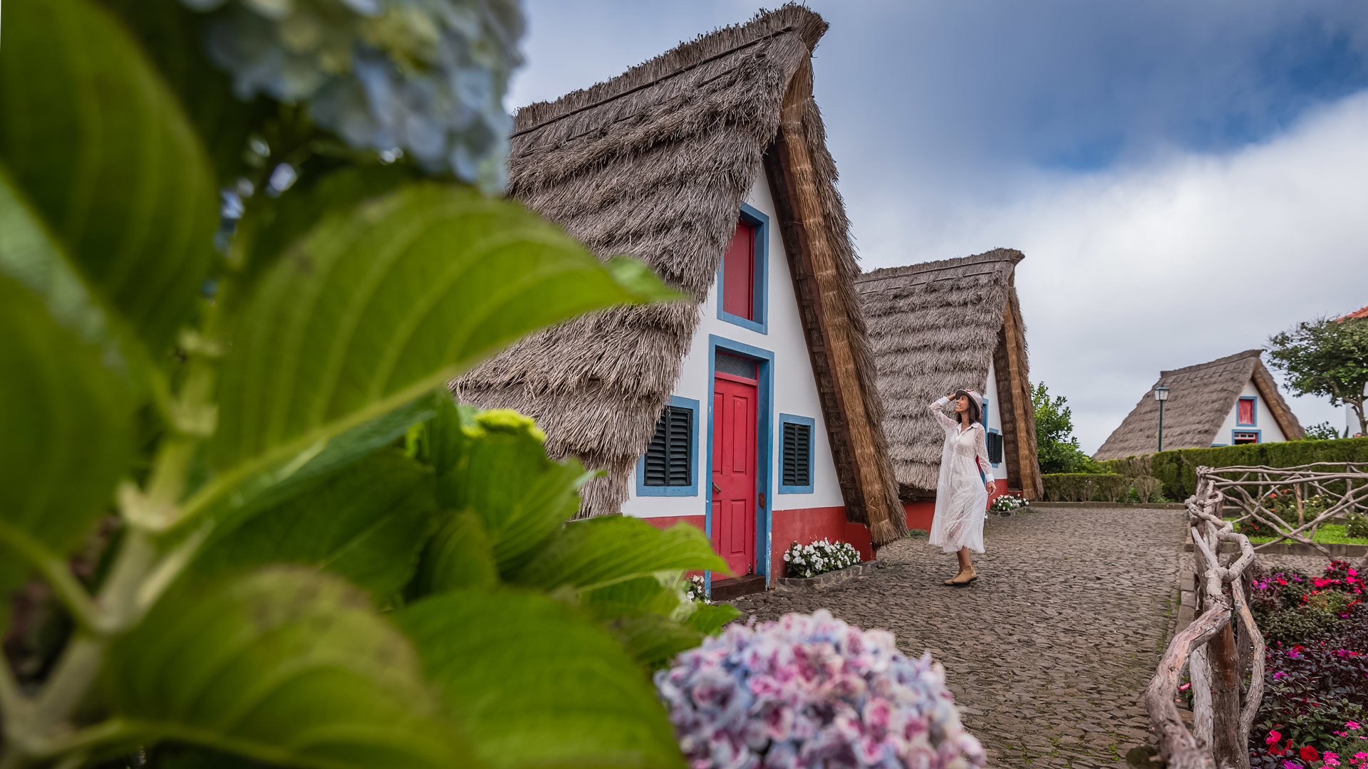Femme en robe blanche près d’une maison triangulaire au toit de chaume.