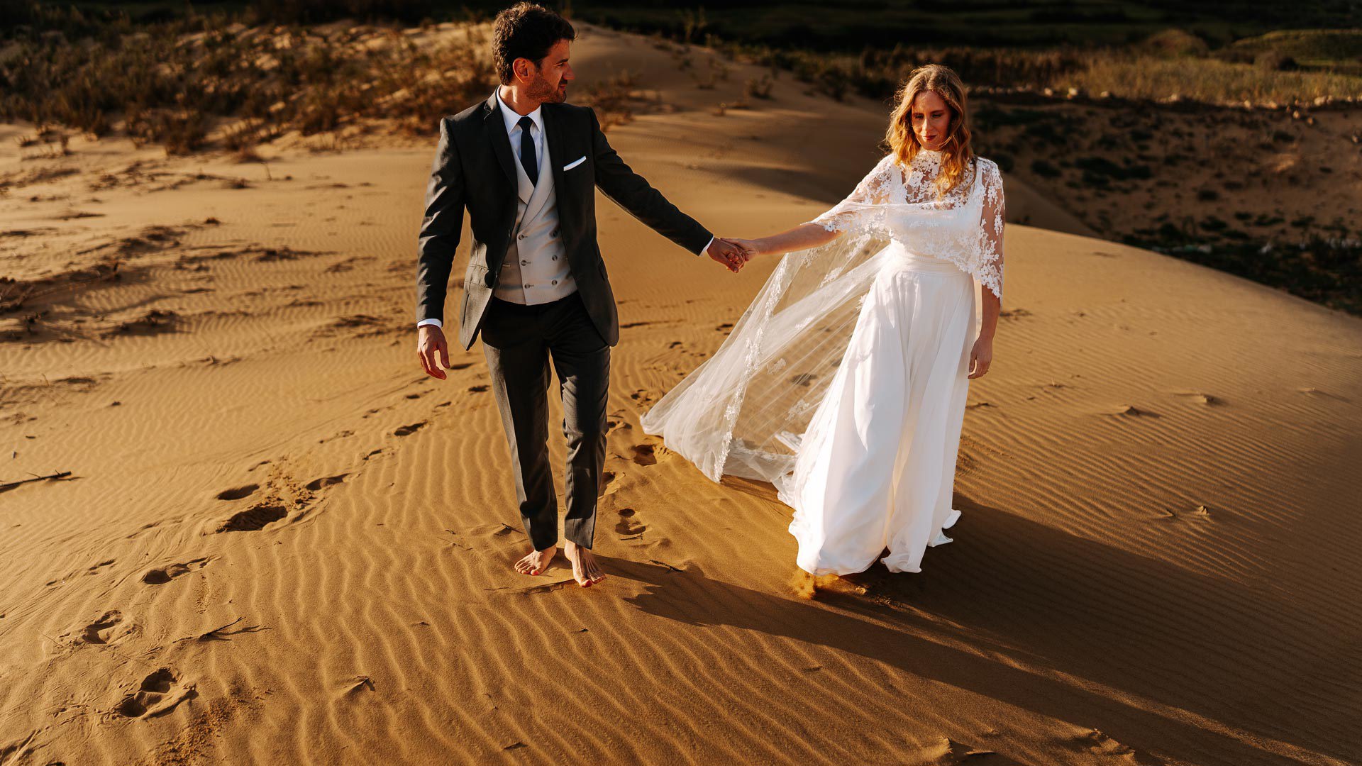 Casal em trajes de casamento a caminhar de mão dada na areia.