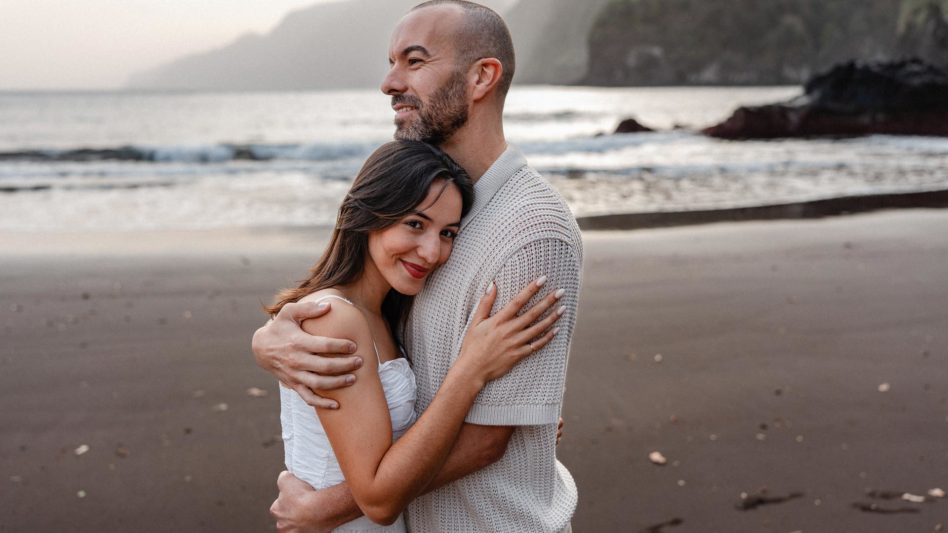 Casal abraçado na praia de areia preta do Seixal, com a costa ao fundo.
