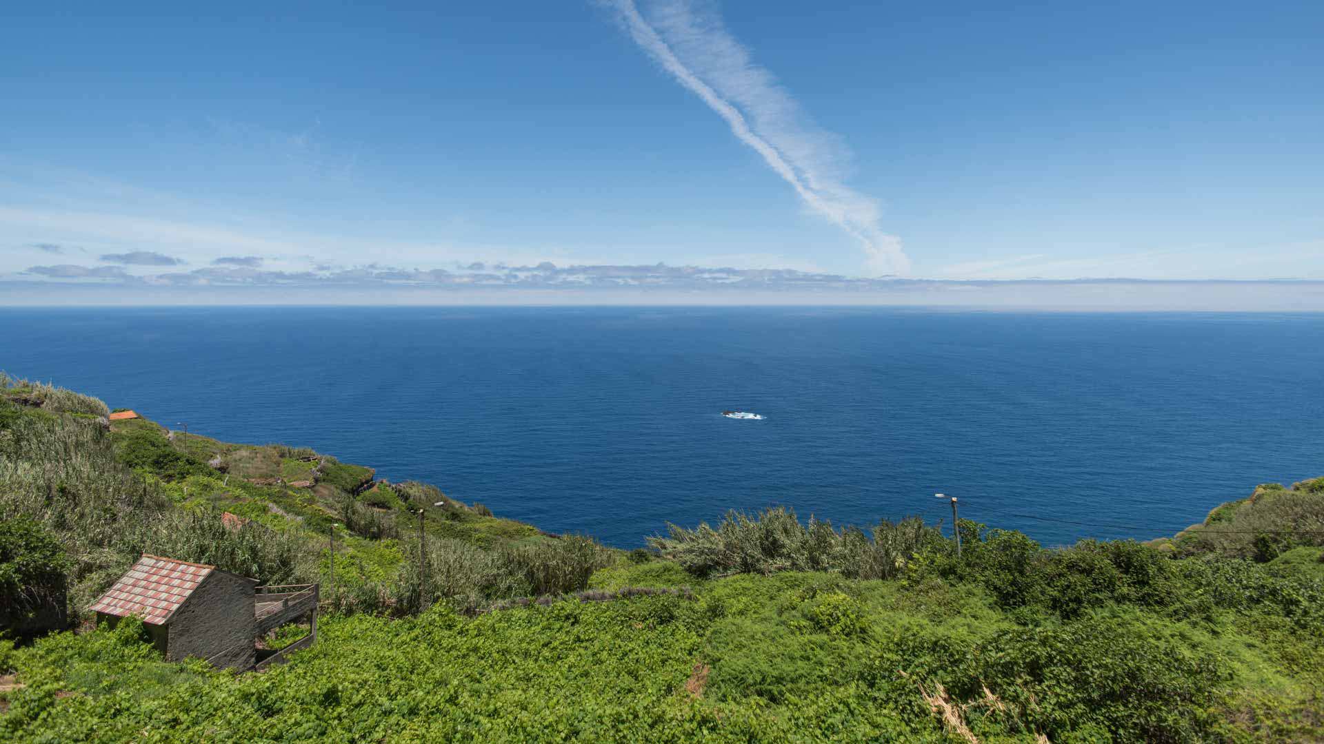 Grüne Hänge mit Steinhaus links und Meer im Hintergrund.