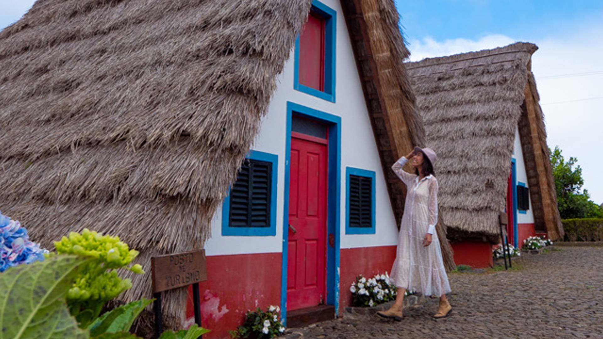 Mujer con vestido blanco y sombrero junto a casa con techo de paja.