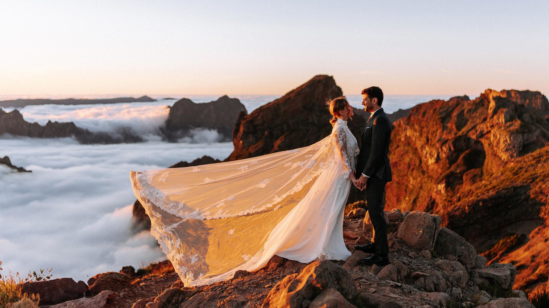 Couple in wedding attire in the mountains above the clouds, watching the sunset.
