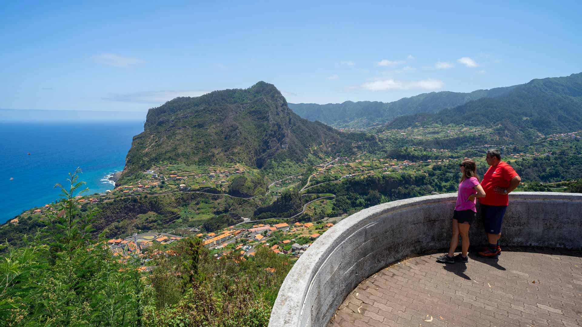 Dos personas en mirador con vista a valle verde y casas.