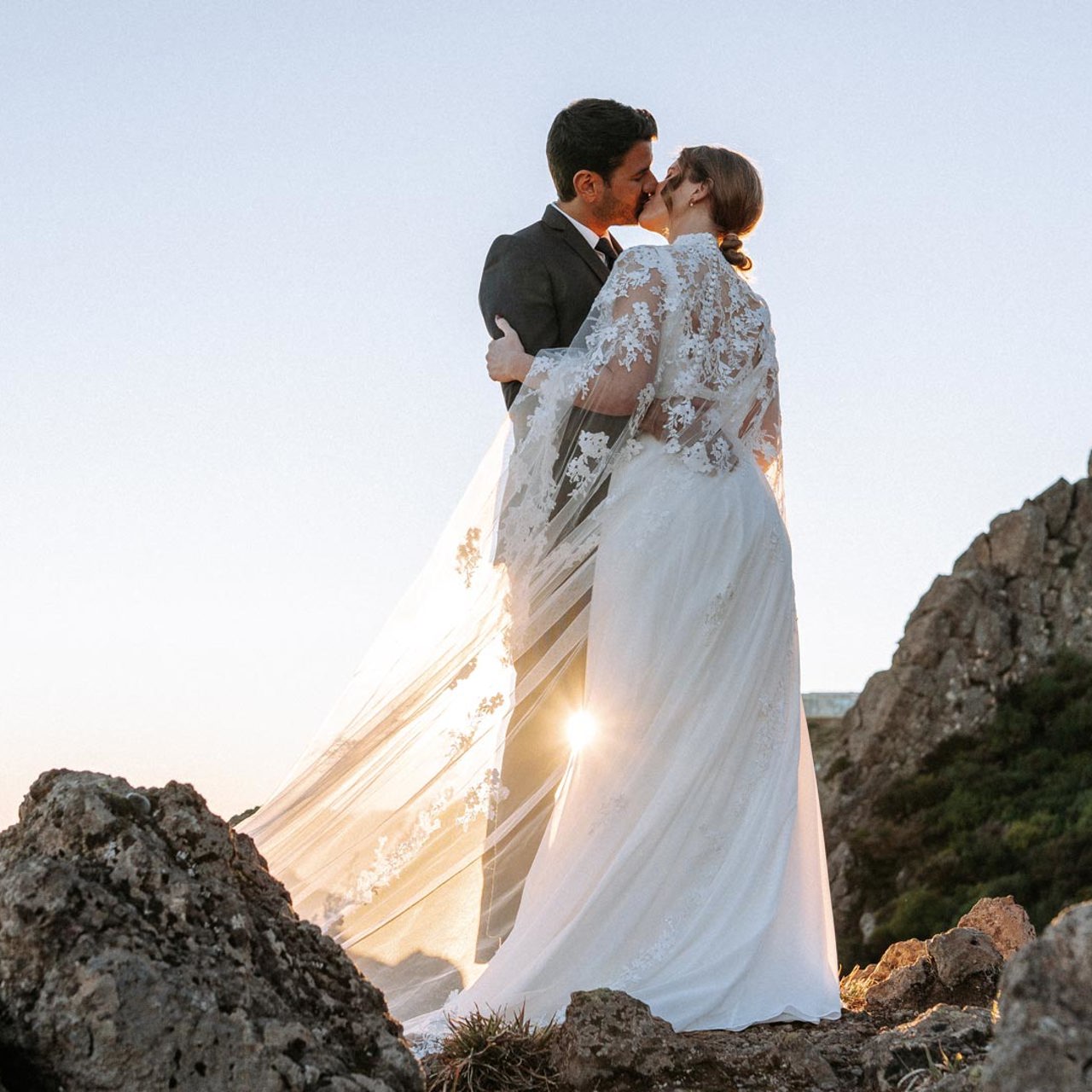 Casal em trajes de casamento nas montanhas, acima das nuvens, a beijar-se.