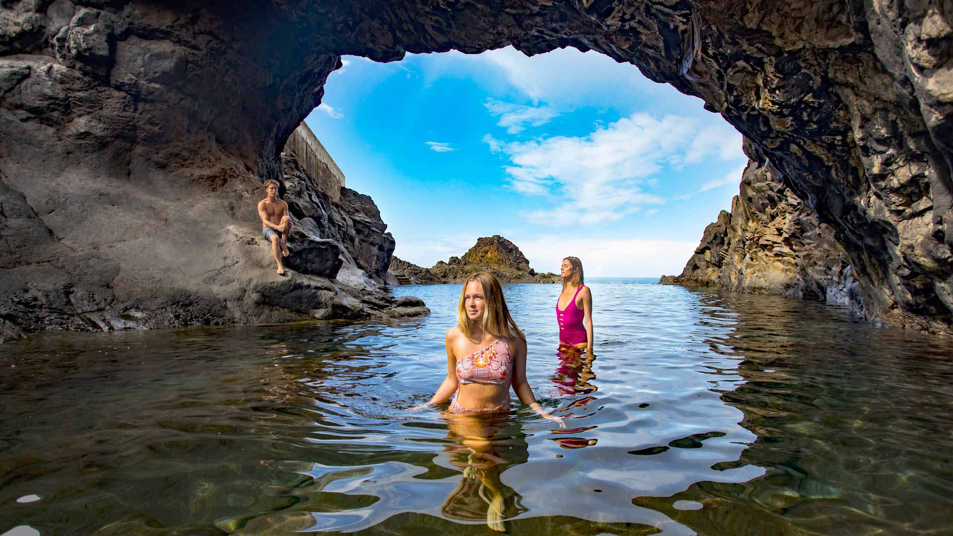 Junge sitzt auf einem Felsen, zwei Mädchen im Wasser des Naturpools auf Madeira.