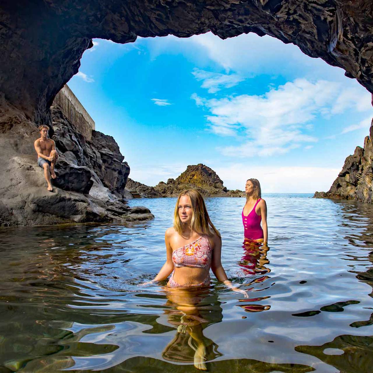 Rapaz sentado na rocha e duas raparigas na água da piscina natural na Madeira.