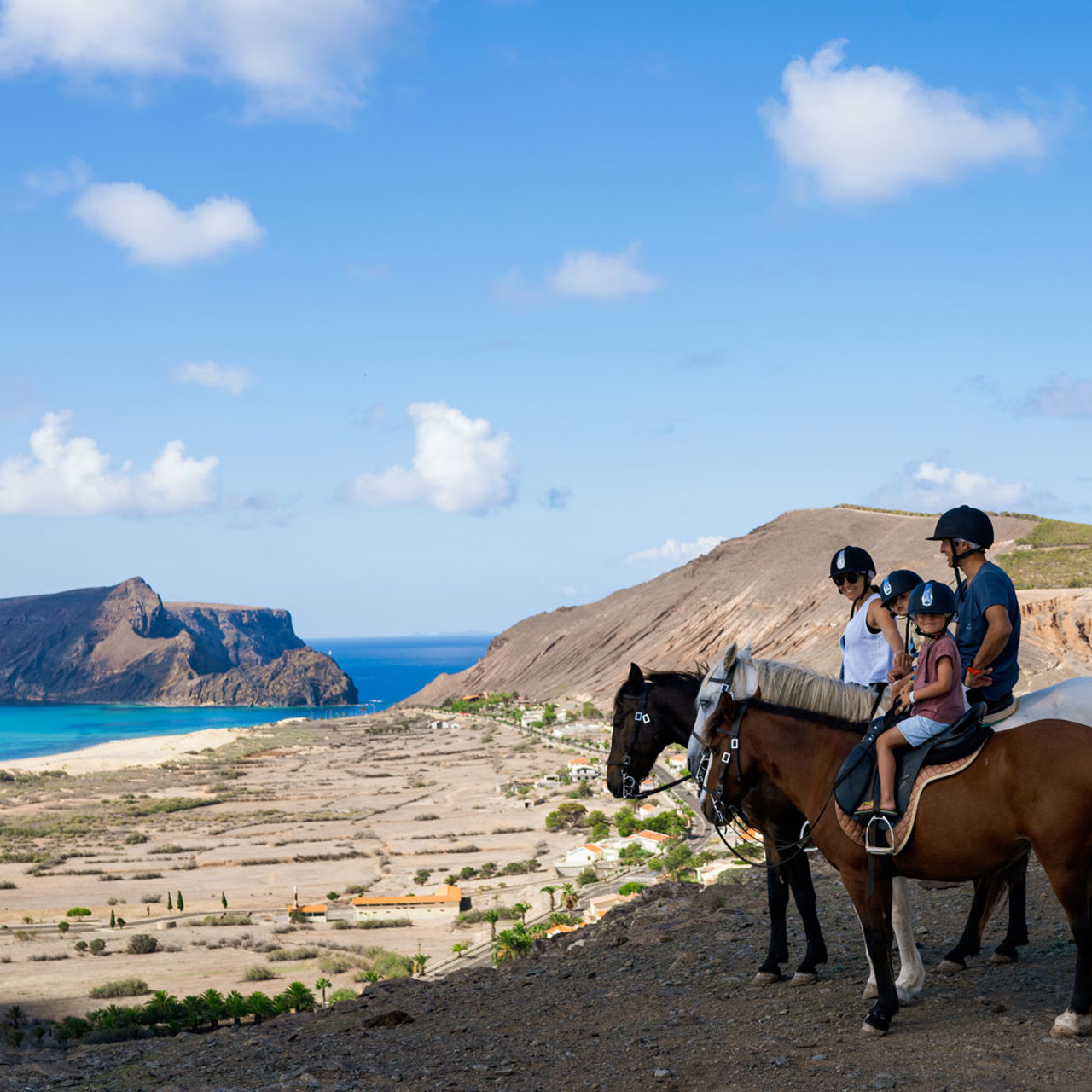 Familia montando 3 caballos por la ladera de Porto Santo, con vista a la playa.