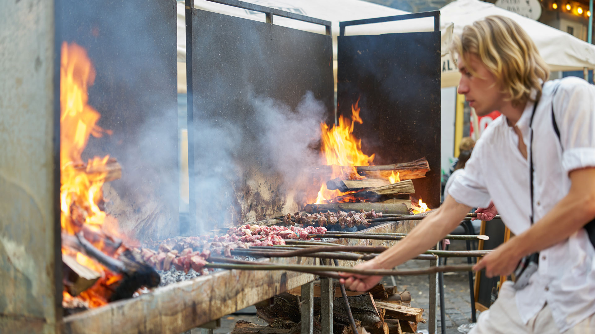 Madeira-Espetada wird über offenem Feuer auf einem Lorbeerholzspieß gegrillt, mit Brennholz im Hintergrund.