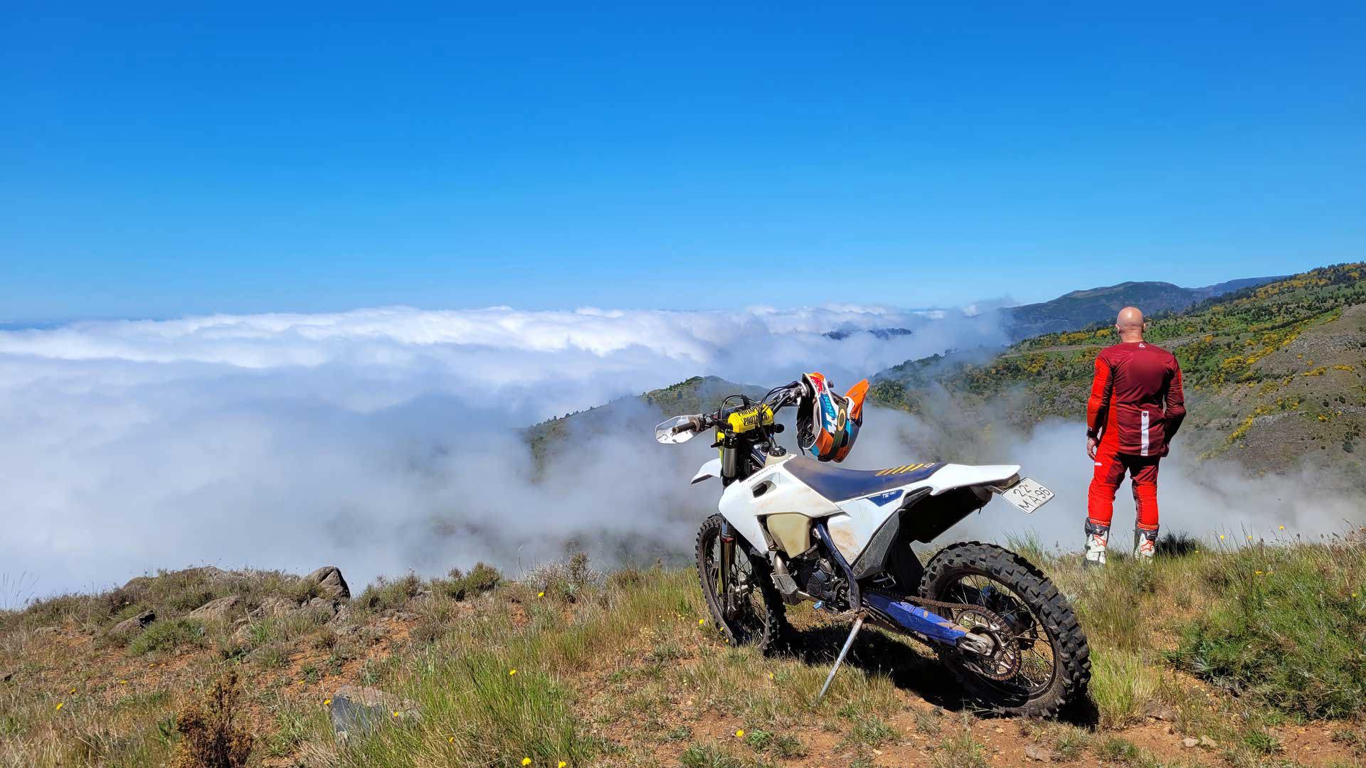 Moto tout-terrain arrêtée avec personne admirant la vue sous les nuages, entourée par la nature.