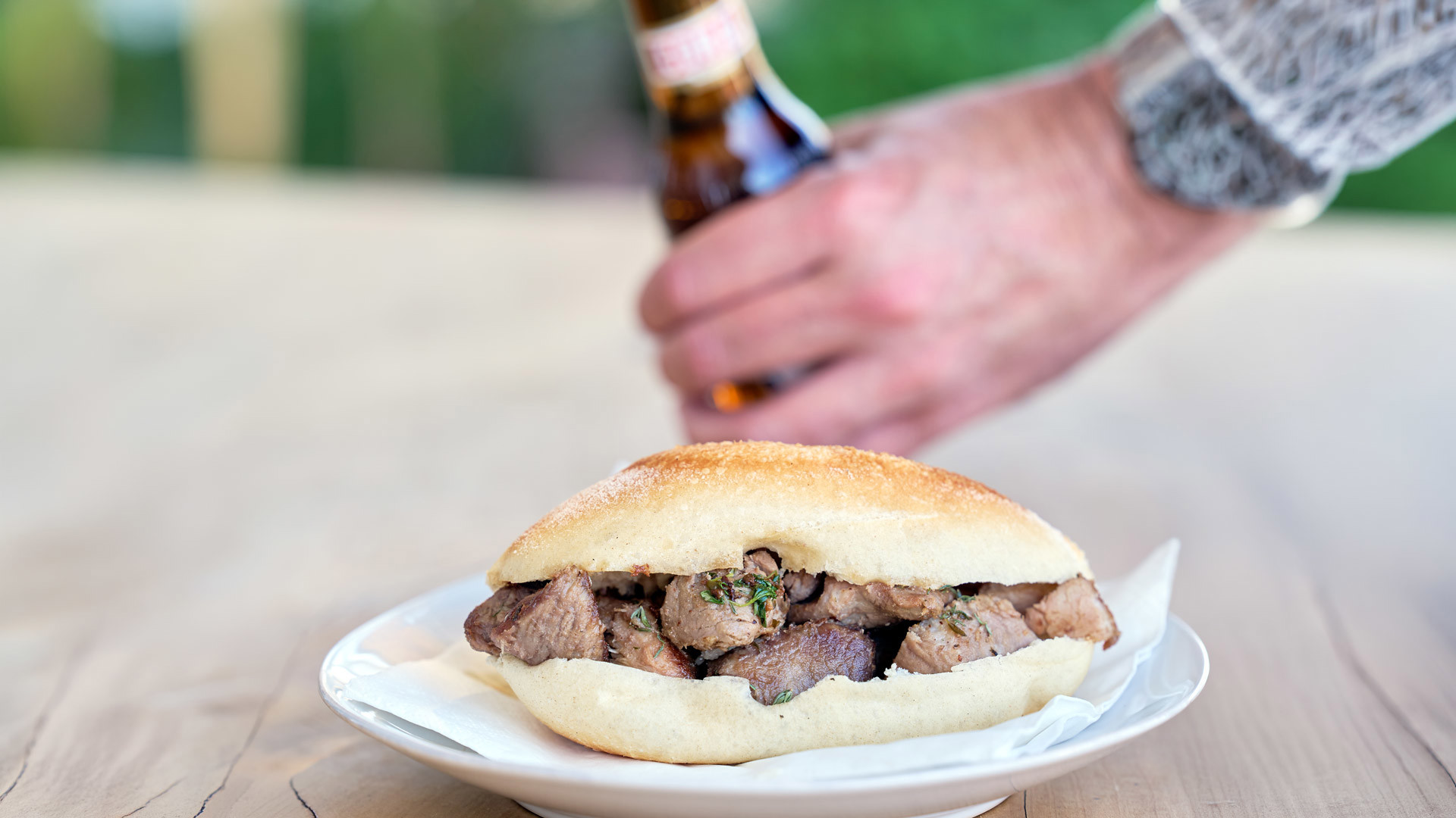 Plate with a carne de vinho e alhos sandwich and a hand holding a beer bottle.