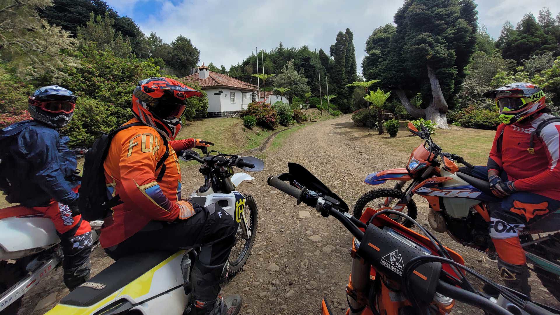 Trois personnes en motos enduro passant devant une maison sur un terrain en terre avec végétation.