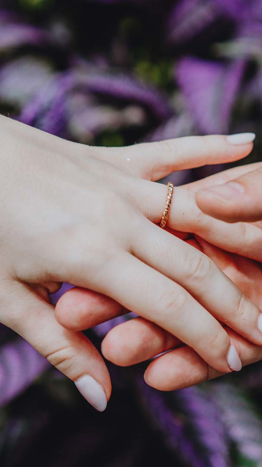 Hände mit Ring, mit lila Blumen im Hintergrund.  