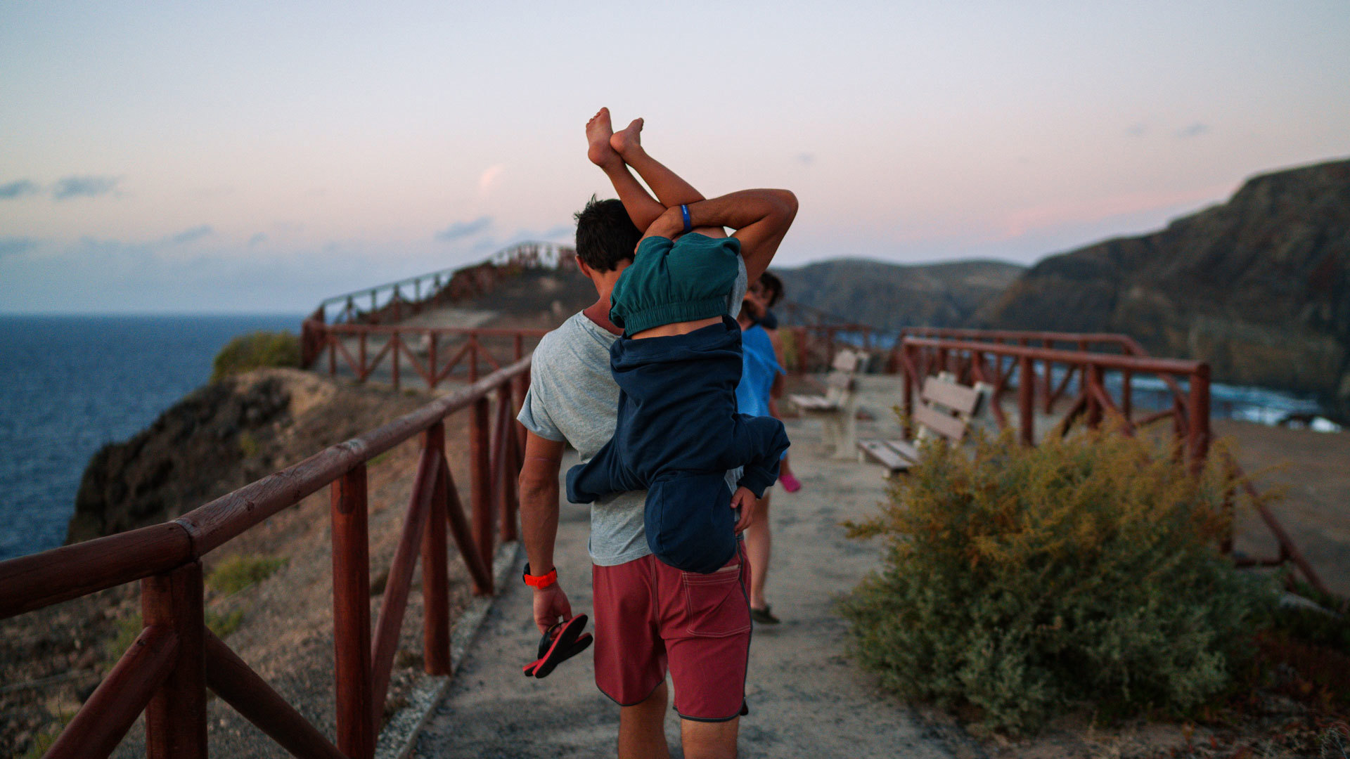 Vater mit Kind auf den Schultern auf dem Strandsteg in Porto Santo bei Sonnenuntergang, mit Hang im Hintergrund.