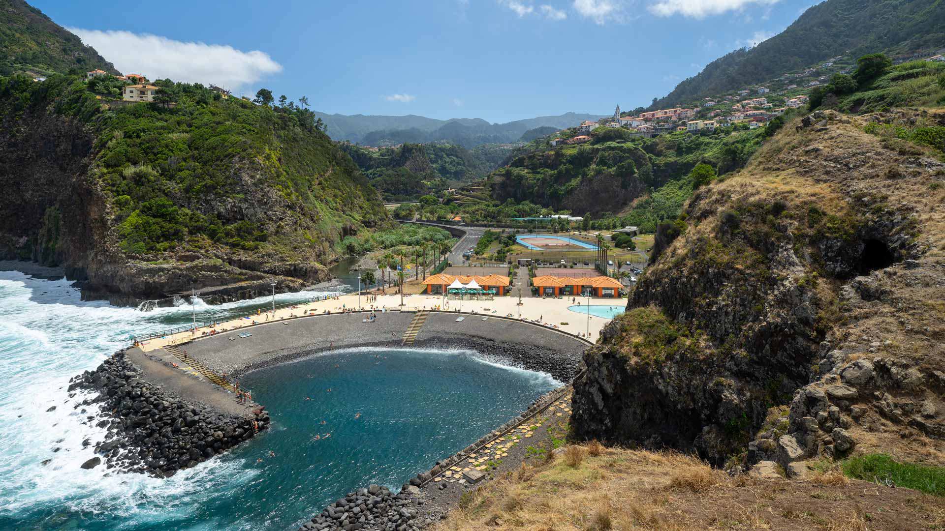 Kieselstrand mit Wellen, Bergen und Häusern im Hintergrund auf Madeira.