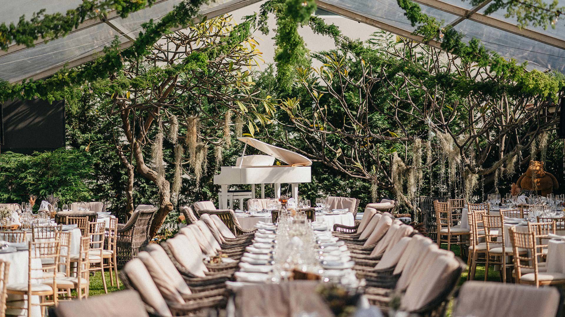 Mesas de jantar decoradas sob tenda com vegetação suspensa, piano e ambiente de casamento.
