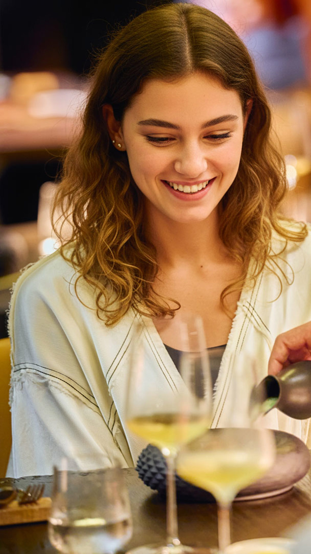 Femme souriante à table en train de savourer un repas, avec une assiette et un verre devant elle.