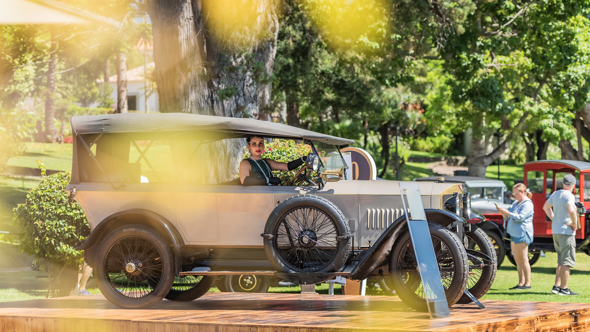 Klassisches Auto in einem Garten ausgestellt, mit Personen drumherum und einer Frau darin sitzend.