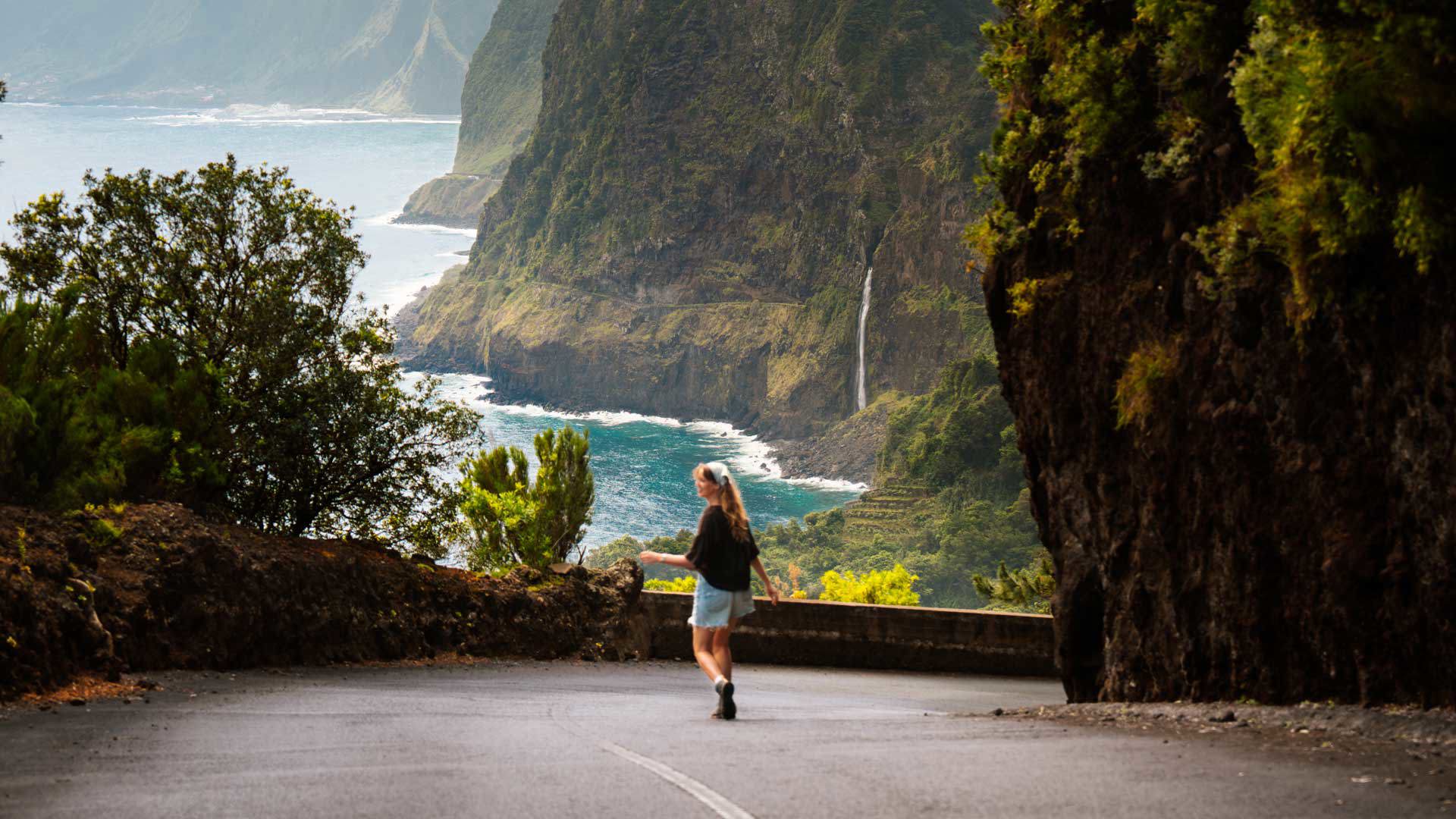 Woman on a road in Madeira with vegetation, waterfall and sea in the background.