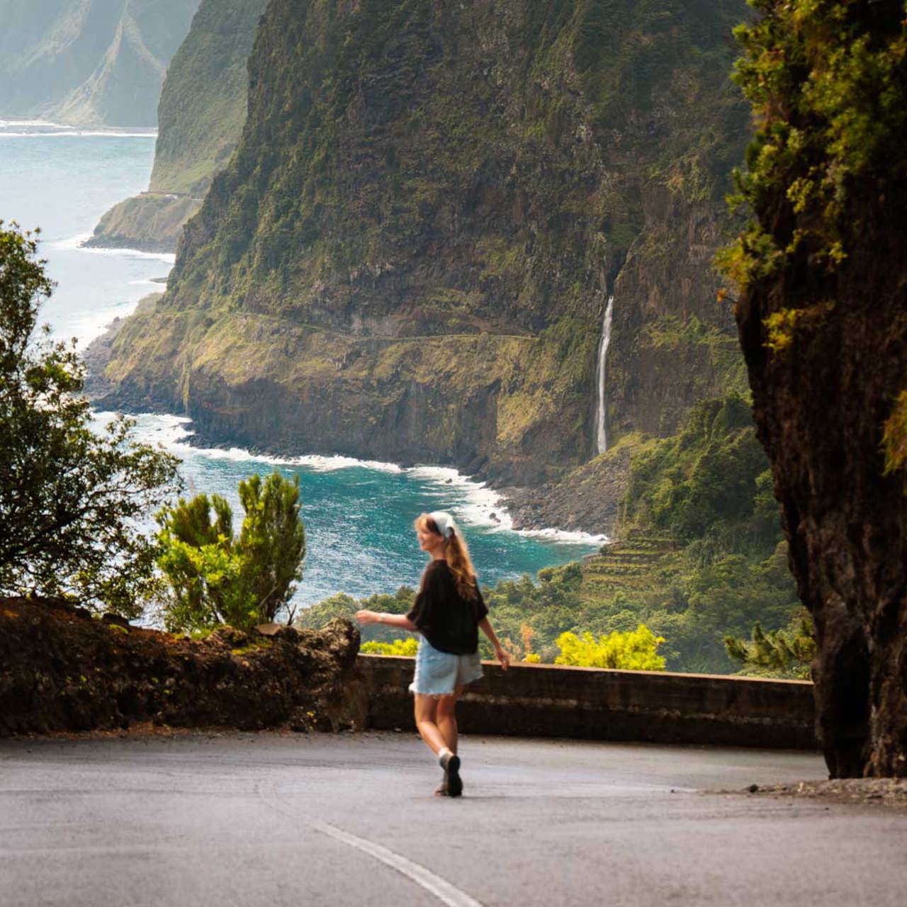 Woman on a road in Madeira with vegetation, waterfall and sea in the background.